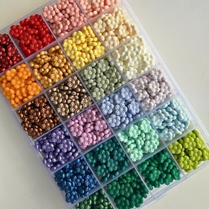 May include: A clear plastic storage box filled with small flower-shaped beads in a rainbow of colors. The beads are arranged in rows, with each row containing a different color.
