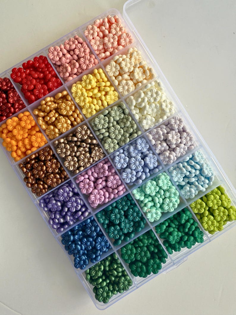 May include: A clear plastic storage box filled with small flower-shaped beads in a rainbow of colors. The beads are arranged in rows, with each row containing a different color.
