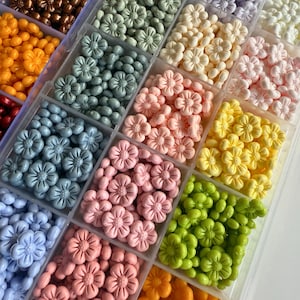 May include: A clear plastic storage box filled with small flower-shaped beads in various colors, including light blue, pink, yellow, green, white, and purple.