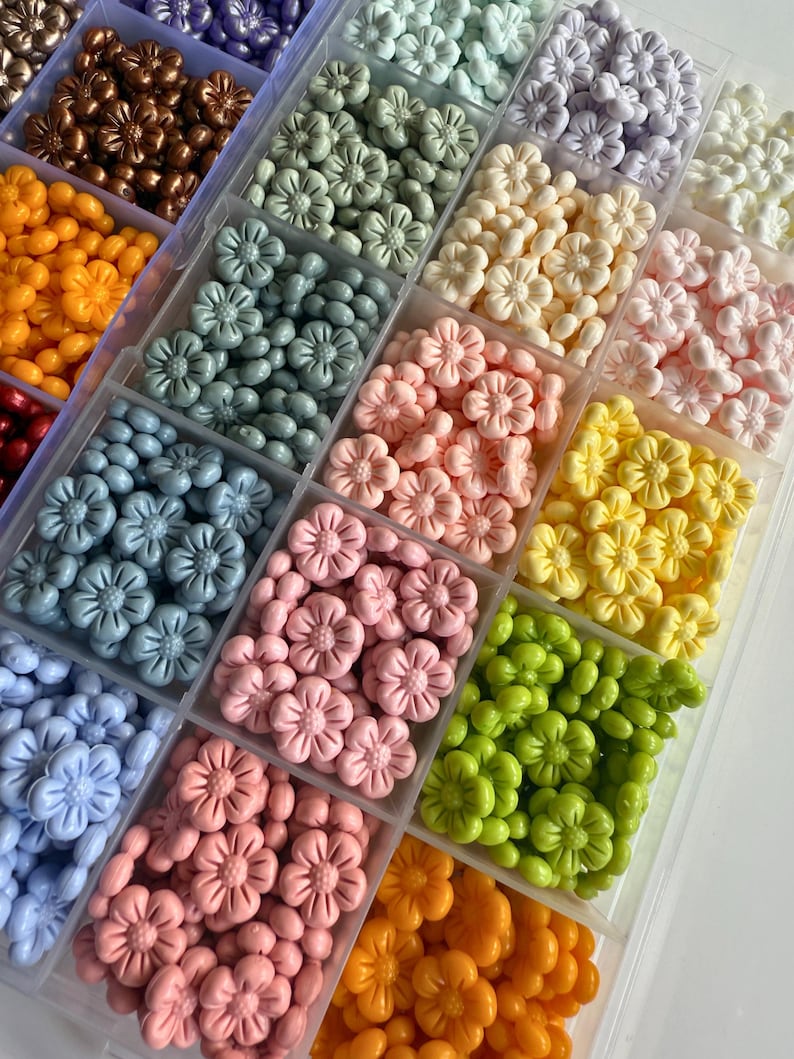 May include: A clear plastic storage box filled with small flower-shaped beads in various colors, including light blue, pink, yellow, green, white, and purple.