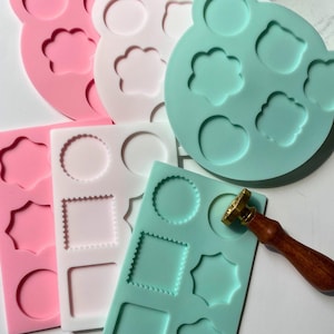 May include: Three silicone molds for crafting, one pink, one white, and one light green. Each mold has different shapes, including circles, squares, stars, and flowers. A gold and wooden wax seal stamp is also included.
