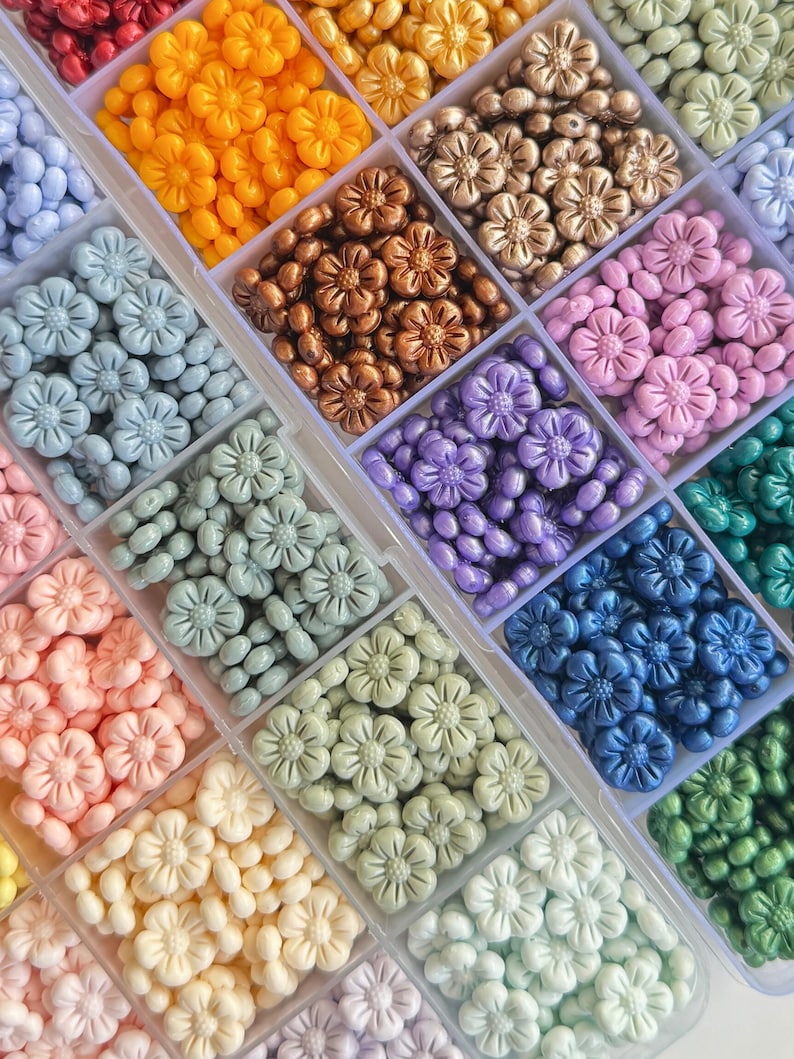 May include: A clear plastic storage box filled with various colors of flower-shaped beads. The beads are arranged in rows and are in shades of red, orange, yellow, brown, purple, blue, green, and white.