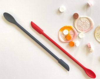 May include: Two silicone spatulas, one red and one gray, with rounded spoon-shaped ends. The spatulas are lying on a white surface with various colored wax seals scattered around them.