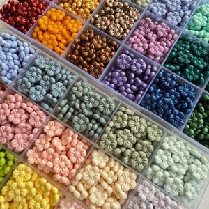 May include: A clear plastic storage box filled with small flower-shaped beads in various colors, including red, orange, yellow, green, blue, purple, pink, and white.