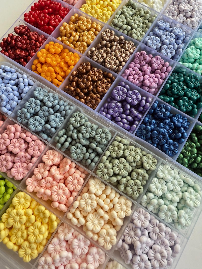 May include: A clear plastic storage box filled with small flower-shaped beads in various colors, including red, orange, yellow, green, blue, purple, pink, and white.