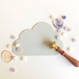 May include: A light gray cloud-shaped silicone mat with a gold and wooden wax seal stamp and several small purple and white wax seals scattered around it. The wax seal stamp has a decorative design on the top. The mat is on a white background. The wax seals say "Thank You".