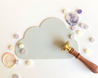 May include: A light gray cloud-shaped silicone mat with a gold and wooden wax seal stamp and several small purple and white wax seals scattered around it. The wax seal stamp has a decorative design on the top. The mat is on a white background. The wax seals say "Thank You".