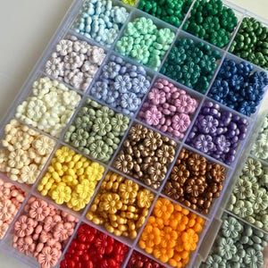 May include: A clear plastic storage box filled with small flower-shaped beads in various colors, including green, blue, purple, white, yellow, orange, red, and brown.