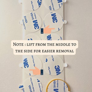 May include: A close-up of several round, white, adhesive 3M double-coated tissue tape circles. The tape is labeled "3M 9448A Double Coated Tissue Tape". The text "NOTE: LIFT FROM THE MIDDLE TO THE SIDE FOR EASIER REMOVAL" is written in black text on a white background.