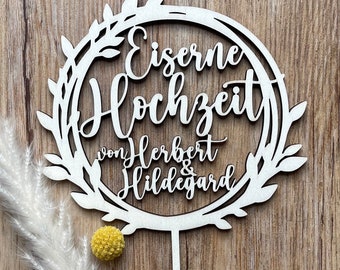 Może przedstawiać: Biały drewniany topper na tort z niemieckim tekstem "Eiserne Hochzeit von Herbert & Hildegard" wewnątrz okrągłego wieńca z liści. Topper ma spiczasty patyczek do włożenia w tort.