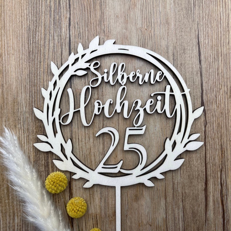 May include: A white wooden cake topper with a wreath design and the text "Silberne Hochzeit 25".