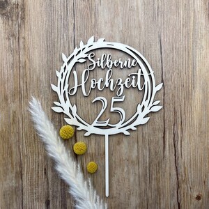 May include: White wooden cake topper with a circle design featuring a leafy wreath and the text "Silberne Hochzeit 25".