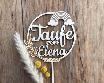 May include: Wooden circle with the text "Taufe von Elena" and the date "02.02.2025". A rainbow with clouds is above the text. The circle is attached to a wooden stick with a small bouquet of yellow flowers.