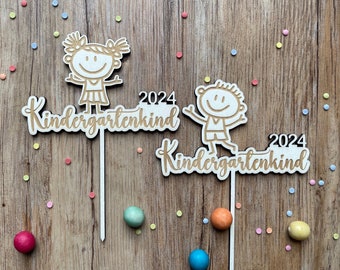 May include: Two wooden cake toppers with the words "Kindergartenkind 2024" and cartoon figures of a girl and a boy. The toppers are on white sticks, with colorful confetti and candies scattered on a wooden surface.