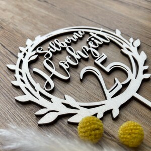May include: A white wooden cake topper with a wreath design and the text "Silberne Hochzeit 25" in cursive.
