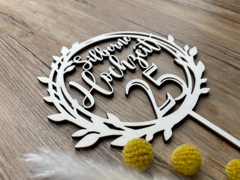 May include: A white wooden cake topper with a wreath design and the text "Silberne Hochzeit 25" in cursive.