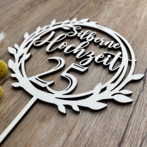 May include: White wooden cake topper with a wreath design and the text "Silberne Hochzeit 25" in a cursive font.