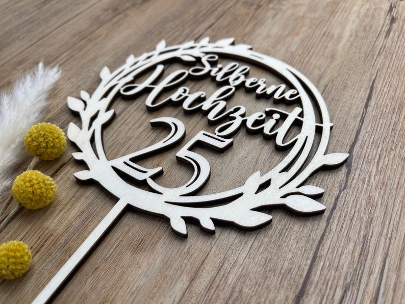 May include: White wooden cake topper with a wreath design and the text "Silberne Hochzeit 25" in a cursive font.