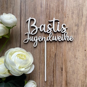 Könnte beinhalten: Weißer Holz-Kuchentopper mit dem Text "Bastis Jugendweihe" in Kursivschrift.
