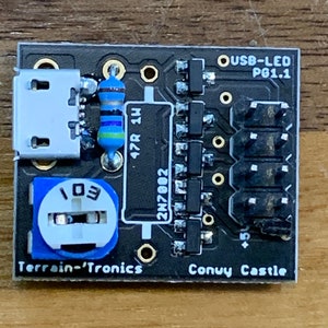 Puede incluir: Una placa de circuito USB-LED PG1.1 negra con varios componentes electrónicos, incluyendo un puerto micro USB, un potenciómetro y varias resistencias. La placa tiene el texto "Terrain-'Tronics" y "Conuy Castle" impreso.