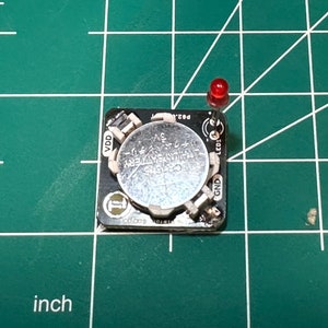 May include: A small black circuit board with a silver CR2032 lithium battery and a red LED. The board has various electronic components and is on a green grid surface. The battery is labeled "CR2032 LITHIUM BATTERY 3V".