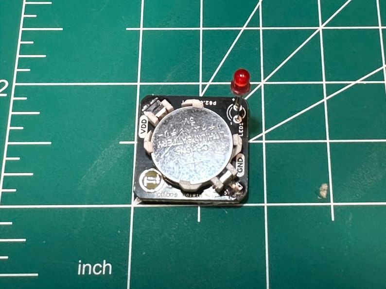 May include: A small black circuit board with a silver CR2032 lithium battery and a red LED. The board has various electronic components and is on a green grid surface. The battery is labeled "CR2032 LITHIUM BATTERY 3V".
