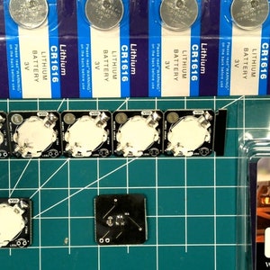 May include: An assortment of electronic components, including CR1616 lithium batteries, small black circuit boards with white circular components, and a package of 10x 3mm flickering LEDs. The items are arranged on a green grid cutting mat.