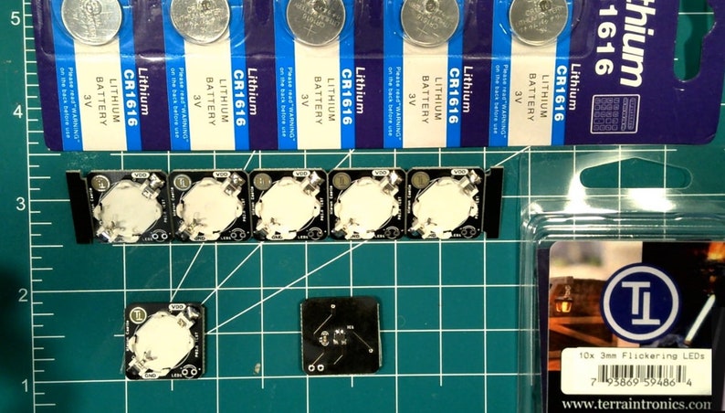 May include: An assortment of electronic components, including CR1616 lithium batteries, small black circuit boards with white circular components, and a package of 10x 3mm flickering LEDs. The items are arranged on a green grid cutting mat.