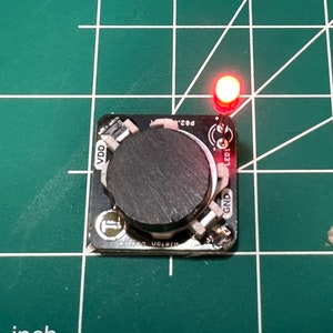 May include: A small black circuit board with a large, dark gray knob in the center. A red LED is lit at the top right. The board is on a green grid surface.
