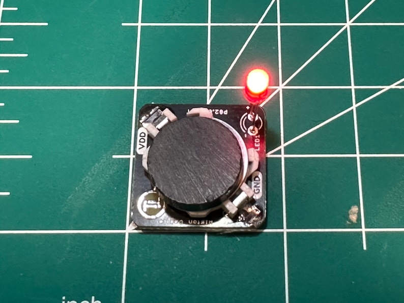 May include: A small black circuit board with a large, dark gray knob in the center. A red LED is lit at the top right. The board is on a green grid surface.