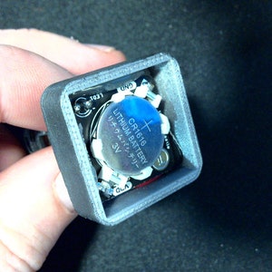 May include: A small, gray, square electronic component with a CR1616 3V lithium battery. The battery is blue and silver, with Japanese text. The component has small circuit board elements and is held in a hand.