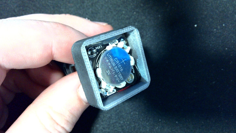 May include: A small, gray, square electronic component with a CR1616 3V lithium battery. The battery is blue and silver, with Japanese text. The component has small circuit board elements and is held in a hand.