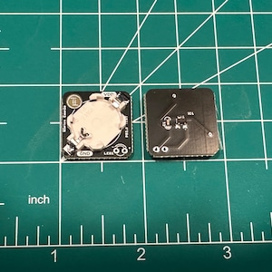 May include: Two small, square black circuit boards with silver components and a battery holder. One board has a battery installed, the other shows the circuit traces. The boards are on a green grid surface, with inch measurements.