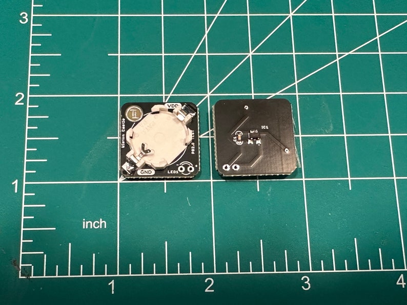 May include: Two small, square black circuit boards with silver components and a battery holder. One board has a battery installed, the other shows the circuit traces. The boards are on a green grid surface, with inch measurements.