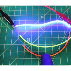 May include: A blue neon light, illuminated and connected to red and black crocodile clips. Below the neon light are three curved wires in yellow and orange. The background is a green cutting mat with grid lines.