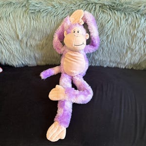 May include: A plush monkey toy featuring long, shaggy purple and white fur with a light beige face, ears, hands, feet, and belly. The monkey has a stitched smile and is posed with its arms raised above its head and one leg bent at the knee.