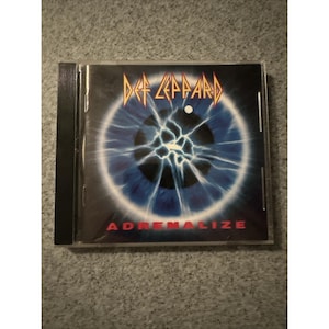 May include: A Def Leppard CD case featuring the album "Adrenalize." The cover art displays a blue eye with a cracked, lightning-like pattern. The band's logo is in a gold, stylized font above the eye.