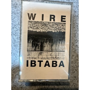 May include: A cassette tape with a white label featuring the band name "WIRE" in bold black letters at the top. Below is the album title "IBTABA" in black. The artwork includes a textured, abstract design in shades of gray and black.