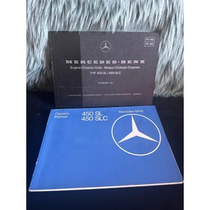 May include: Two Mercedes-Benz manuals. The top manual is black with white text and the Mercedes-Benz logo. The bottom manual is blue with white text and the Mercedes-Benz logo. The manuals are for the 450 SL/450 SLC models.