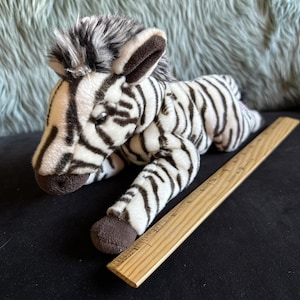 May include: A plush zebra toy with black and white stripes, brown ears, and a gray mane. The toy is lying down next to a wooden ruler. The zebra is a soft, cuddly toy.