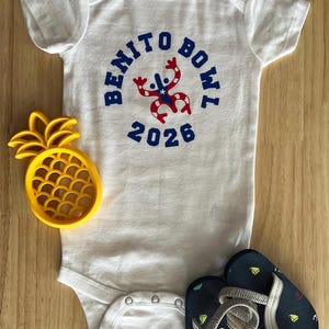 Op de afbeelding: Witte babybody met de tekst "BENITO BOWL 2026" in blauw, met een rood-blauwe afbeelding. Een geel ananasvormig speelgoed en marineblauwe teenslippers met anker- en zeilbootontwerpen zijn ook in de afbeelding.