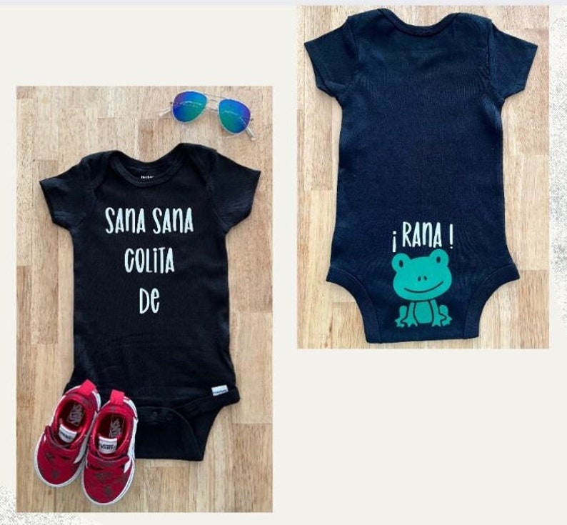 May include: Two black baby onesies. One onesie has the text "Sana Sana Colita De" printed in white on the front. The other onesie has a green frog with the text "&iexcl;Rana!" printed in white on the back.