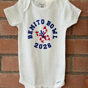 Op de afbeelding: Witte babybody met de tekst "BENITO BOWL 2026" in blauw. Het ontwerp bevat een rood, wit en blauw grafisch element. De body hangt voor een bakstenen muur.