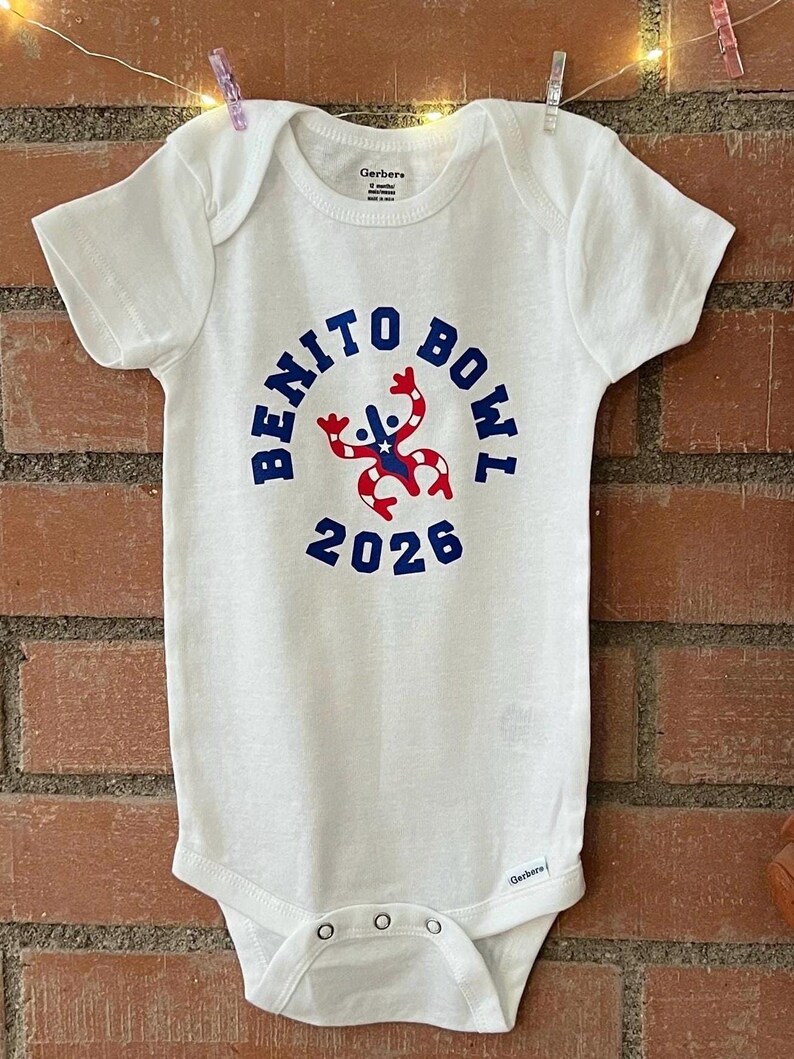 Op de afbeelding: Witte babybody met de tekst "BENITO BOWL 2026" in blauw. Het ontwerp bevat een rood, wit en blauw grafisch element. De body hangt voor een bakstenen muur.