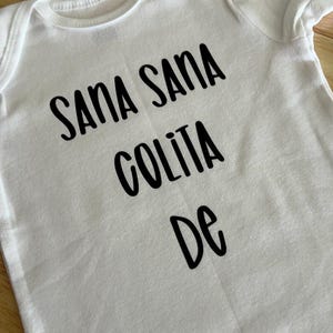 May include: White baby onesie with black text that reads "Sana Sana Colita De".