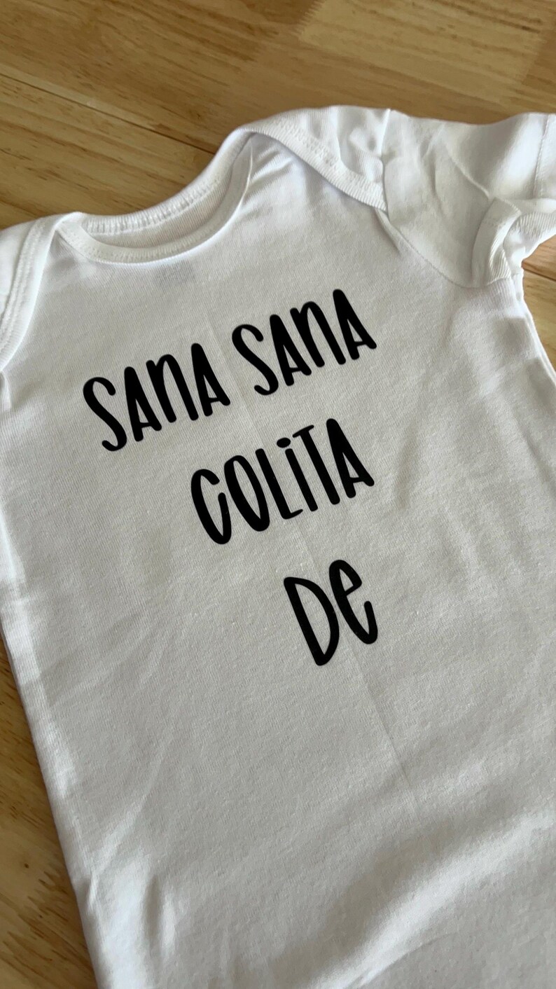 May include: White baby onesie with black text that reads "Sana Sana Colita De".