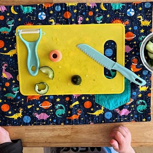 May include: A yellow cutting board with a blue knife, a green vegetable peeler, and a few slices of cucumber on a blue and black fabric placemat with a dinosaur pattern.