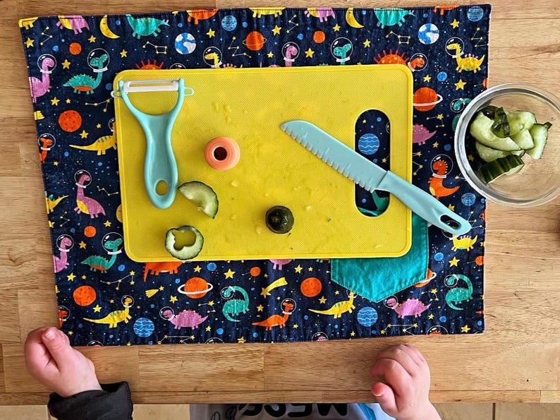 May include: A yellow cutting board with a blue knife, a green vegetable peeler, and a few slices of cucumber on a blue and black fabric placemat with a dinosaur pattern.