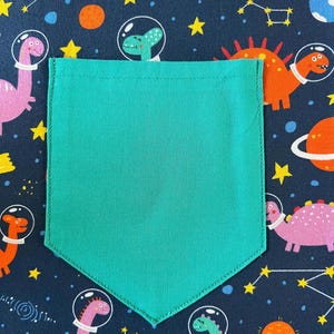 May include: A turquoise fabric pocket with a stitched edge, against a dark blue background with a repeating pattern of dinosaurs and stars.