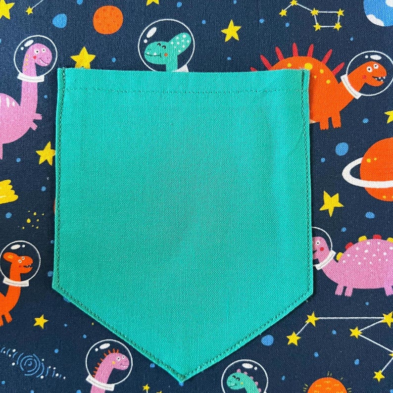 May include: A turquoise fabric pocket with a stitched edge, against a dark blue background with a repeating pattern of dinosaurs and stars.